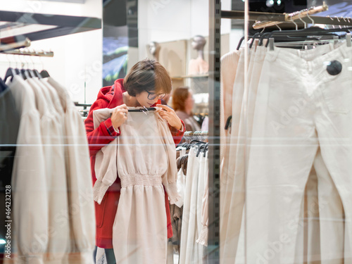 Woman chooses dress at clothing store. Casual trousers hanging on hangers. View through transparent shop window. Shopping at mall. Basic clothes for everyday wear. Modern urban fashion.