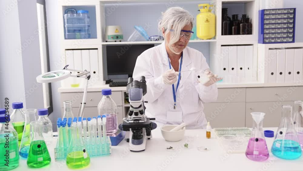 Middle age grey-haired woman wearing scientist uniform mixing liquid working at laboratory Middle age grey-haired woman wearing scientist uniform mixing liquid working at laboratory