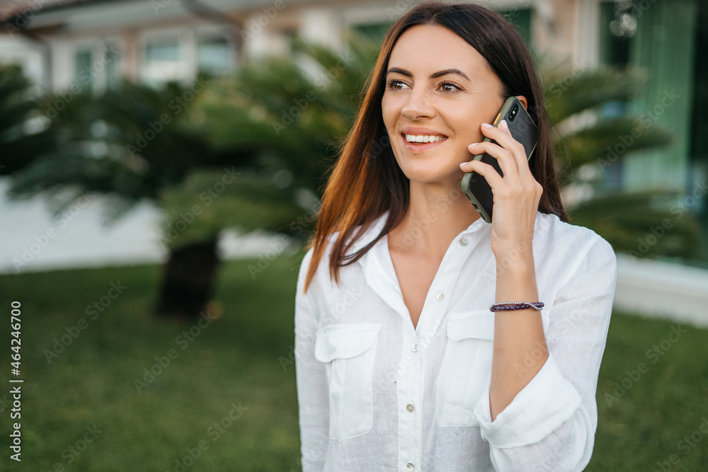Portrait of Attractive happy i brunette woman smiling using smart phone in summer garden detailing in white shirt. The concept of communication and comfort. Portrait of Attractive happy i brunette woman smiling using smart phone in summer garden detailing in white shirt. The concept of communication and comfort.