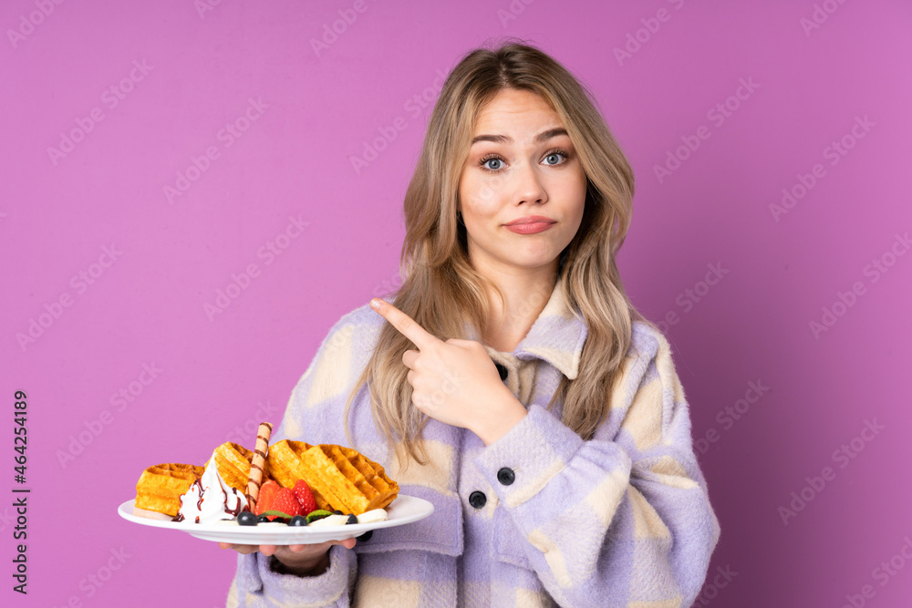 Teenager Russian girl holding waffles isolated on purple background pointing to the laterals having doubts Teenager Russian girl holding waffles isolated on purple background pointing to the laterals having doubts