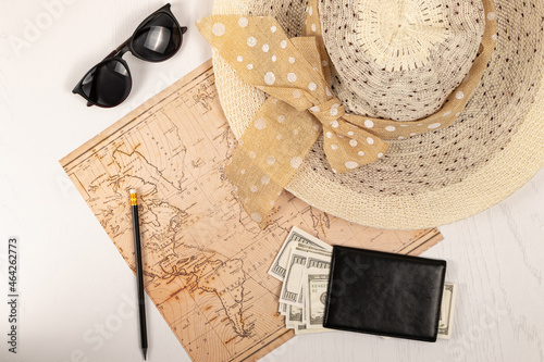world map, straw hat, black leather wallet with money, sunglasses and a pencil on a white wooden table. view from above