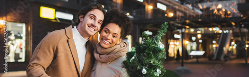 happy multiethnic couple holding small christmas pine in evening, banner.