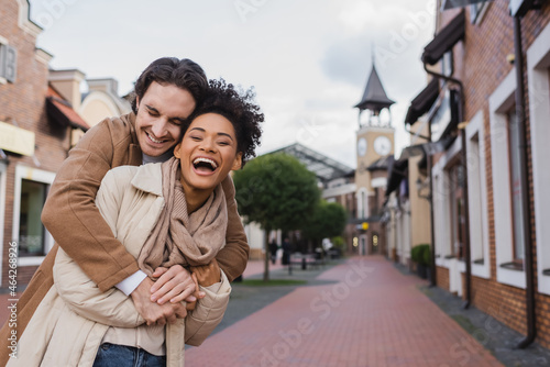 happy man smiling while hugging amazed african american girlfriend outdoors.