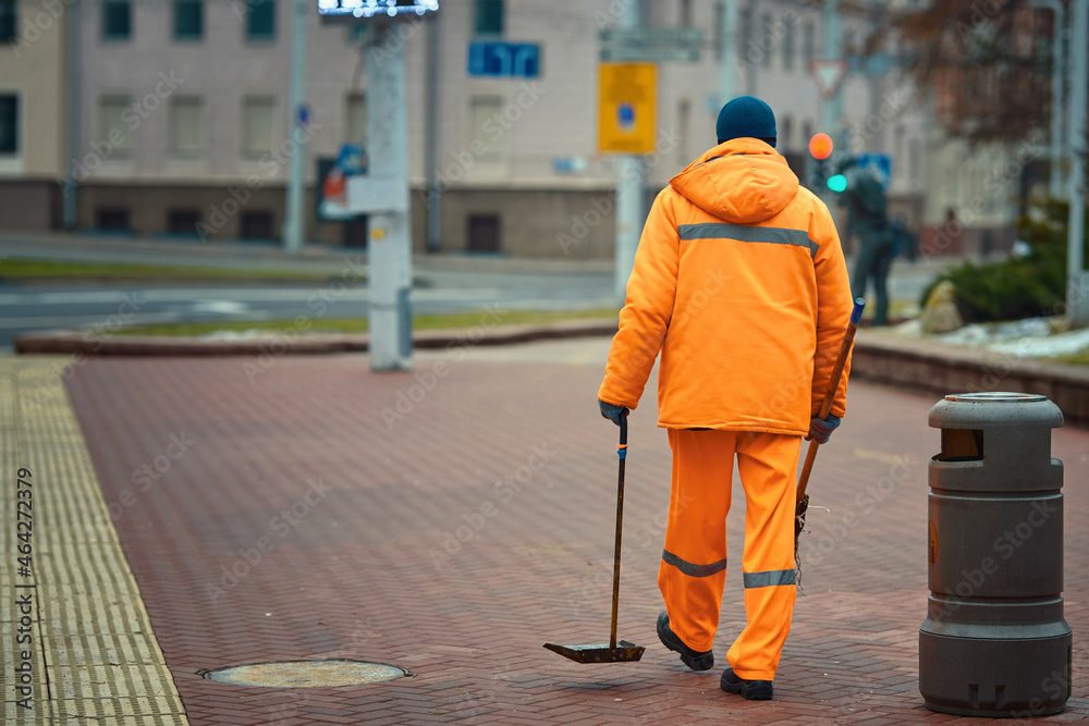 Street sweaper cleanup road and pedestrian zone in the city. Municipal worker sweep city street, janitor with broomstick and scoop for garbage. Men in orange uniform collecting garbage in the evening.