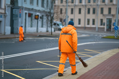Street sweaper team cleanup road and pedestrian zone in the city. Municipal workers sweep city street, janitors with broomstick and scoop for garbage. Men in orange uniform collecting garbage