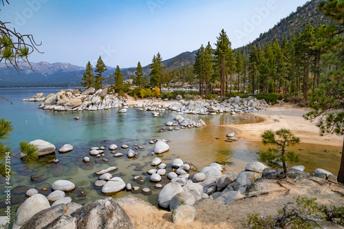 Very rocky shoreline of Lake Tahoe with clear, still blue water, mountains and trees against a blue sky