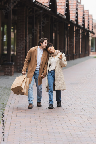 loving multiethnic couple in coats holding hands while walking with purchases near mall. © LIGHTFIELD STUDIOS