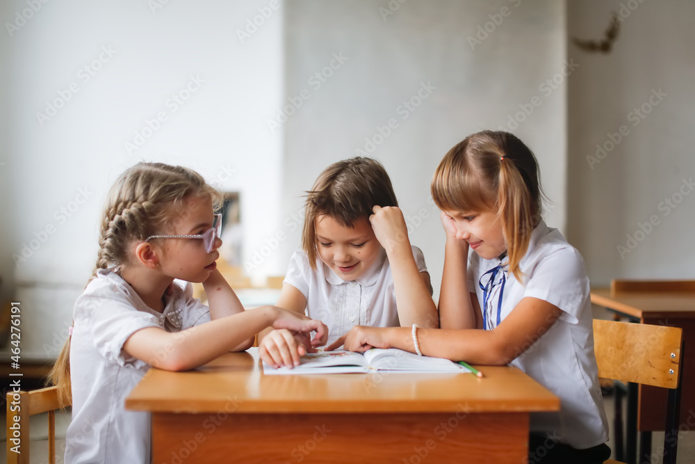 Three girlfriends laughing, classmates read a book together, solve problems. Mutual assistance at school, children sit studying at desks, school learning concept Three girlfriends laughing, classmates read a book together, solve problems. Mutual assistance at school, children sit studying at desks, school learning concept