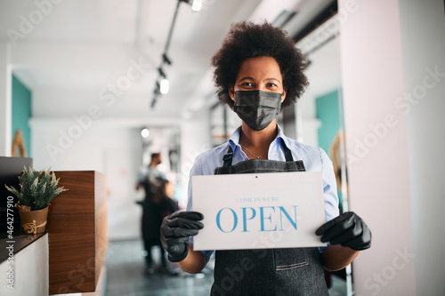 Happy African American hairstylist holds open sign at her salon and wears face mask due to COVID-19 pandemic. photo