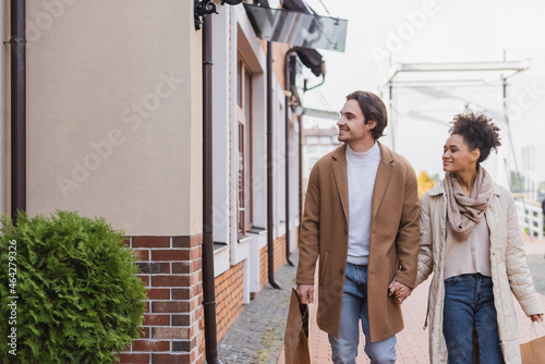 cheerful african american woman holding hands with boyfriend holding shopping bags while walking near mall.