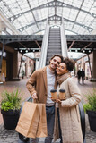 happy african american woman holding coffee to go near man with shopping bags smiling in mall.