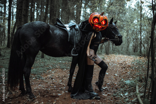 mask in the form of a pumpkin with burning eyes. creepy girl costume for halloween. wrap yourself in a long black cloak. the rider stands next to the black horse