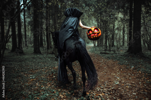 the rider is sitting astride a horse. a girl in a long black raincoat with a pumpkin mask with burning eyes. Creepy cosplay shooting for Halloween in the autumn forest. a cloak fluttering in the wind