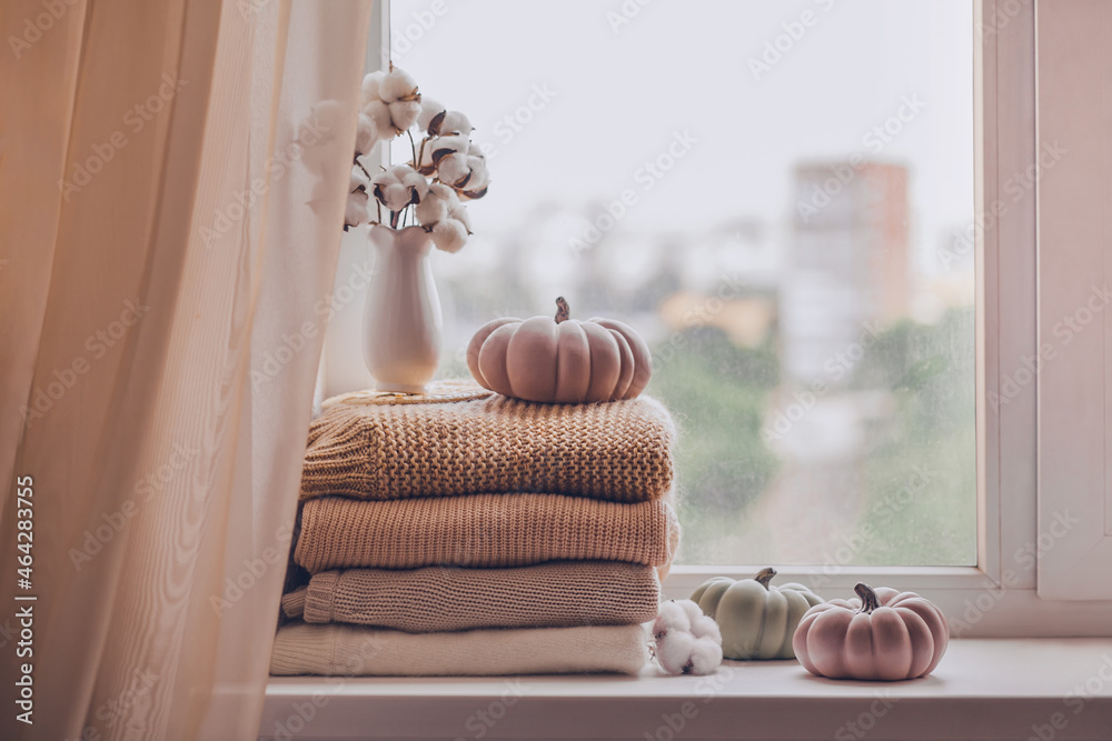 A stack of warm knitted sweaters on the windowsill with pumpkins and a vase of cotton plant in pastel shades