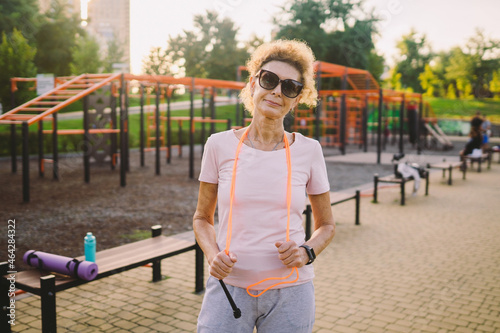 Sporty mature woman posing with a skipping rope in an outdoor sports field. Senior woman holding skipping rope, portrait against the background of the street gym. Active senior lifestyle photo