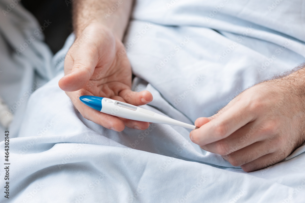 A man lying in bed, holding a thermometer in his hands. Close-up of the hands. The concept of measuring body temperature A man lying in bed, holding a thermometer in his hands. Close-up of the hands. The concept of measuring body temperature