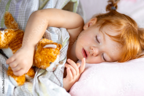 a little red-haired girl sleeping peacefully in her bed and hugs a plush kitten. 