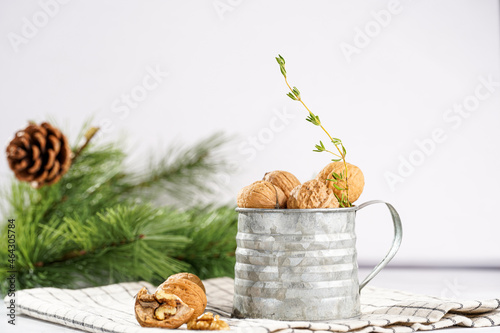 A metal mug with whole brown walnuts on a checkered linen cloth and a christmas fair-tree branch on a white table surface