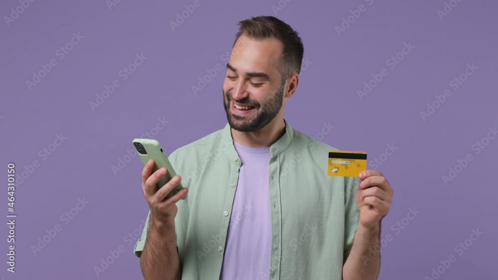 Smiling cheerful young bearded man 20s wears mint shirt using mobile cell phone hold in hand credit bank card doing online shopping crossed isolated on plain light purple background studio portrait Smiling cheerful young bearded man 20s wears mint shirt using mobile cell phone hold in hand credit bank card doing online shopping crossed isolated on plain light purple background studio portrait