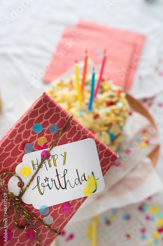 Closeup view of a colorful tagged birthday present tied with a golden ribbon and a blurred cake as background.