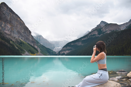 Young girl sitting on rocks taking pictures of lake and mountains with retro film camera