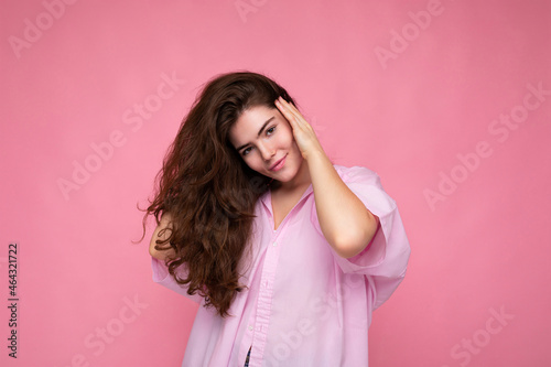 Beautiful charming fascinating young curly brunette woman wearing pink shirt and grey hat isolated on pink background with copy space