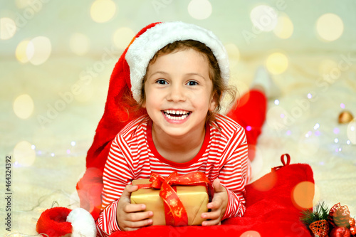 A little smiling girl in Santa's red hat and red christmas clothing lies on the bed with a gift. Preparation for the celebration. New Year. hildren's Christmas. Atmosphere. Home. photo