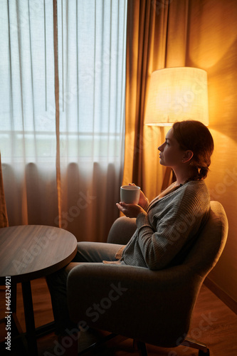 A young woman in home clothes sits in a room near a window with a cup of coffee. It's early morning. The room is lit by the soft light of a floor lamp. Comfort, comfort, hotel, quarantine.