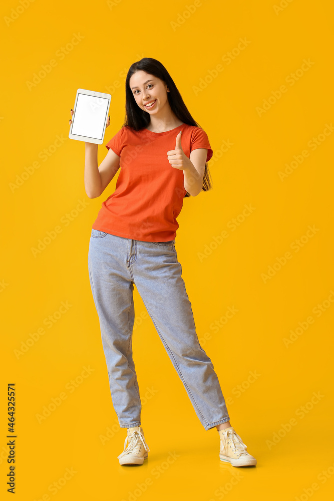 Beautiful teenage girl with tablet computer showing thumb-up on yellow background