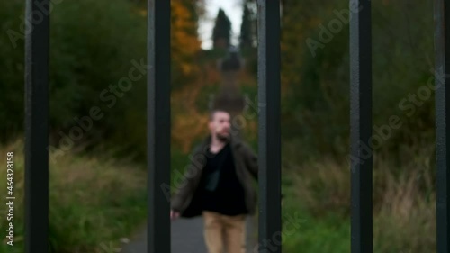 A man runs along the path being deffenetly scared by something or someone then hits enclosure bars with his hands trying to escape yelling for help of fear. a closed green area with grass and trees