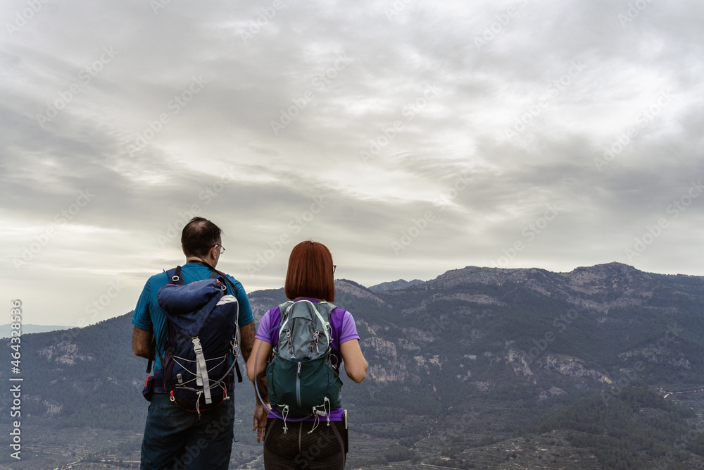 Man and woman hikers with their backs to the camera in the foreground with a mountainous landscape in the background on a cloudy day.
