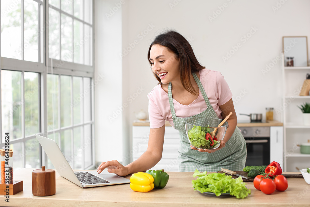Young woman with fresh vegetable salad and laptop in kitchen Young woman with fresh vegetable salad and laptop in kitchen