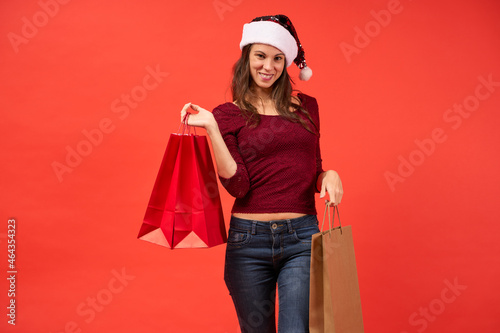 Girl in Santa Claus hat smiling with Christmas shopping bags on orange background