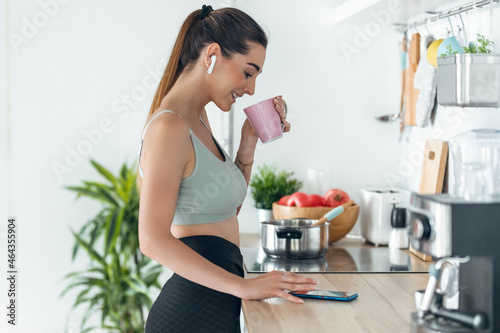 Fototapeta Naklejka Na Ścianę i Meble -  Sporty woman listening to music with her smartphone whiledrinking coffee in the kitchen at home.