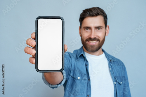 Satisfied confident smiling adult businessman showing mobile phone blank white screen mockup to camera standing isolated on blue background. Man holding smartphone and shows copy space for advertising