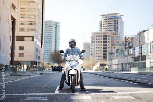 Wide shot of a young motorcyclist stopped at a traffic light in Barcelona. The man riding his scooter through the city on a large avenue lined with skyscrapers photo