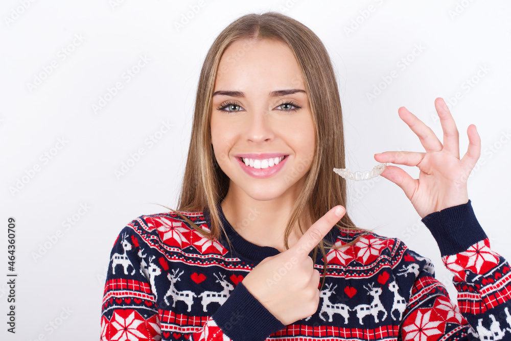 Young beautiful caucasian girl wearing christmas sweaters on white background holding an invisible aligner and pointing at it. Dental healthcare and confidence concept. Young beautiful caucasian girl wearing christmas sweaters on white background holding an invisible aligner and pointing at it. Dental healthcare and confidence concept.