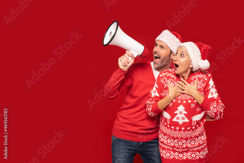 Happy beautiful mature couple in christmas sweaters and santa claus hats shouting on megaphone.