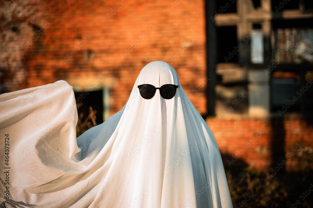 A man in a ghost costume made from a sheet and sunglasses stands near an abandoned building and waves his hand. Ghost Challenge 2021. Spooky season. Celebrating halloween. A man in a ghost costume made from a sheet and sunglasses stands near an abandoned building and waves his hand. Ghost Challenge 2021. Spooky season. Celebrating halloween.