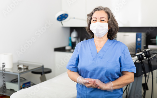 Portrait of an experienced female doctor in a protective mask standing in the clinic office near the workplace