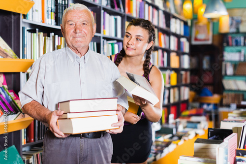 Old man with granddaughter are choosing books in bookstore.