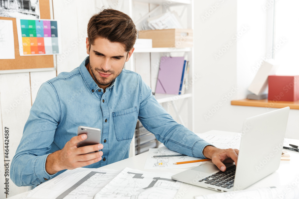 Young man using cellphone while working with laptop and drawings in office