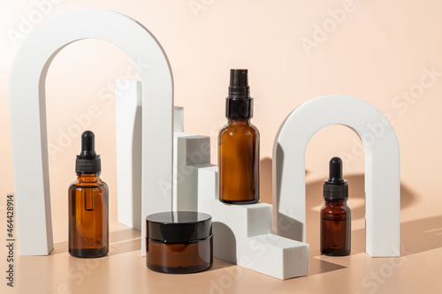 Amber glass dropper bottles and cream jar on white podiums. Beige background. Container mockup with cosmetic oil or serum. Showcase for the presentation skincare products.