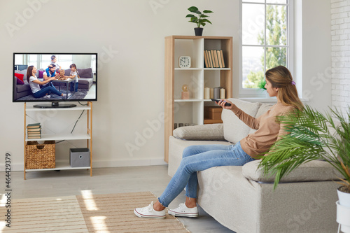 Young woman renter sit rest on couch in living room browse TV program with remote control. Millennial female tenant relax on sofa enjoy television in modern home. Rental, real estate concept.