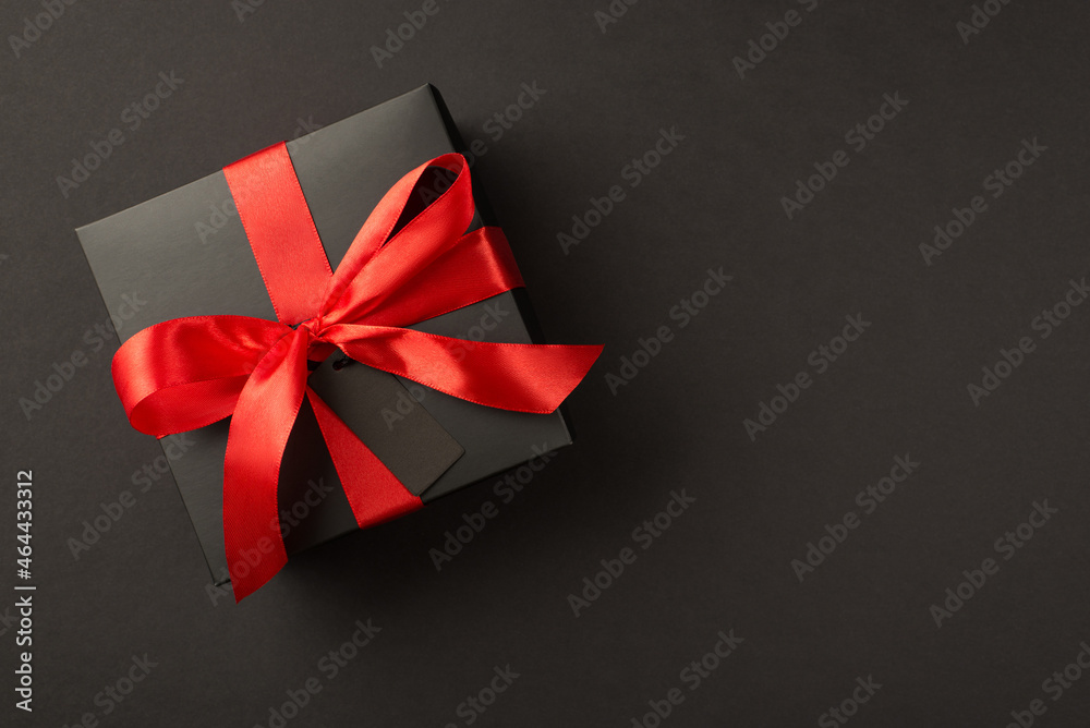 Top view photo of stylish giftbox in black packaging with vivid red satin ribbon bow and tag on isolated black background with copyspace