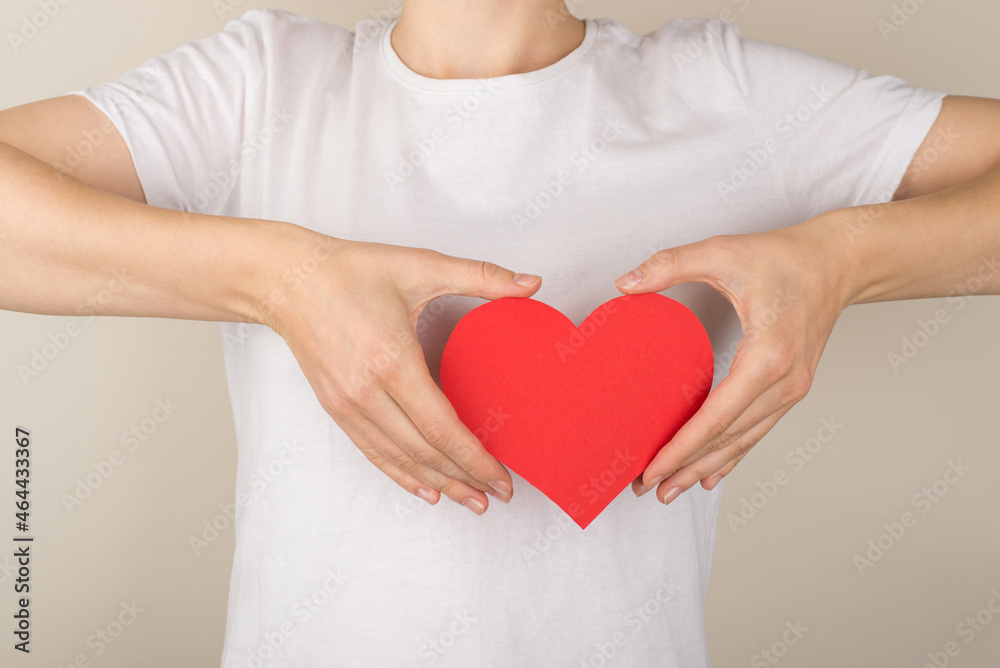 Cropped closeup photo of young woman in white t-shirt holding red paper heart near breast on isolated grey background Cropped closeup photo of young woman in white t-shirt holding red paper heart near breast on isolated grey background