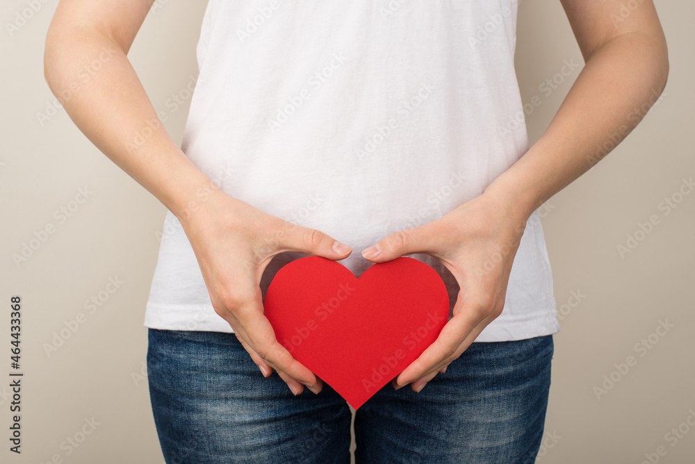 Cropped closeup photo of girl in white t-shirt and jeans holding red paper heart near crotch on isolated grey background Cropped closeup photo of girl in white t-shirt and jeans holding red paper heart near crotch on isolated grey background
