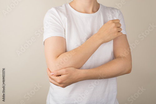 Cropped closeup photo of young woman in white t-shirt holding her right elbow on isolated grey background