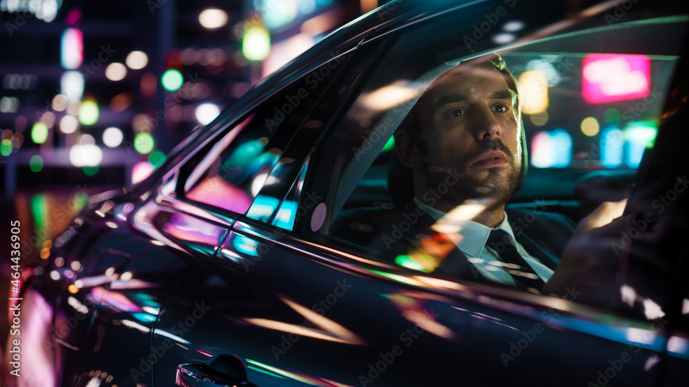 Handsome Businessman in a Suit Commuting from Office in a Backseat of His Car at Night. Entrepreneur Using Smartphone while in Transfer Taxi in Urban City Street with Working Neon Signs. Handsome Businessman in a Suit Commuting from Office in a Backseat of His Car at Night. Entrepreneur Using Smartphone while in Transfer Taxi in Urban City Street with Working Neon Signs.