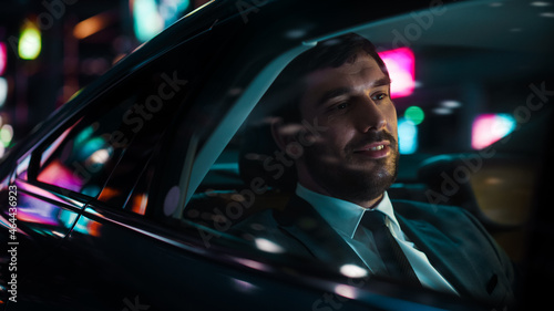Handsome Businessman in a Suit Commuting from Office in a Backseat of His Car at Night. Entrepreneur Passenger Traveling in a Transfer Taxi in Urban City Street with Working Neon Signs.